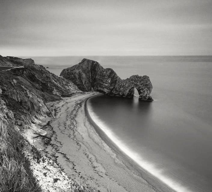 A rock arch standing in a calm sea beside a curved beach