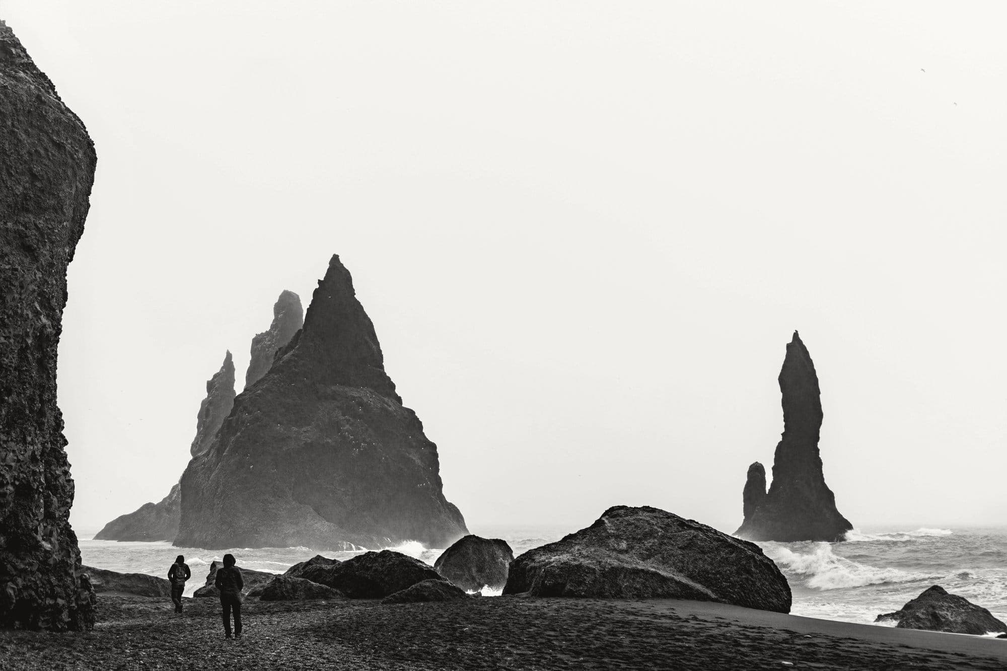 Two figures walking on a black sand beach beneath sea stacks